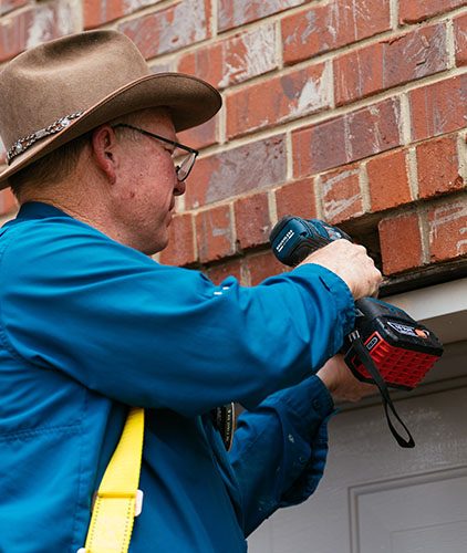 Man wearing a western hat and blue shirt using tool to fix masonry above garage