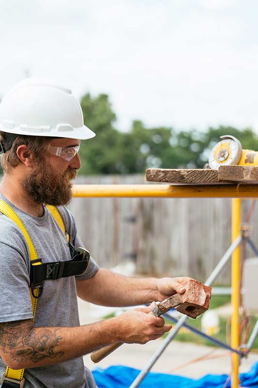 Man with hard hat and beard wearing yellow harness working on bricks
