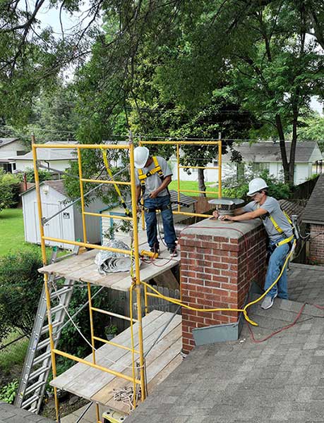 Two men standing on a lifted platform and roof installing a new chimney cap on a brick chimney